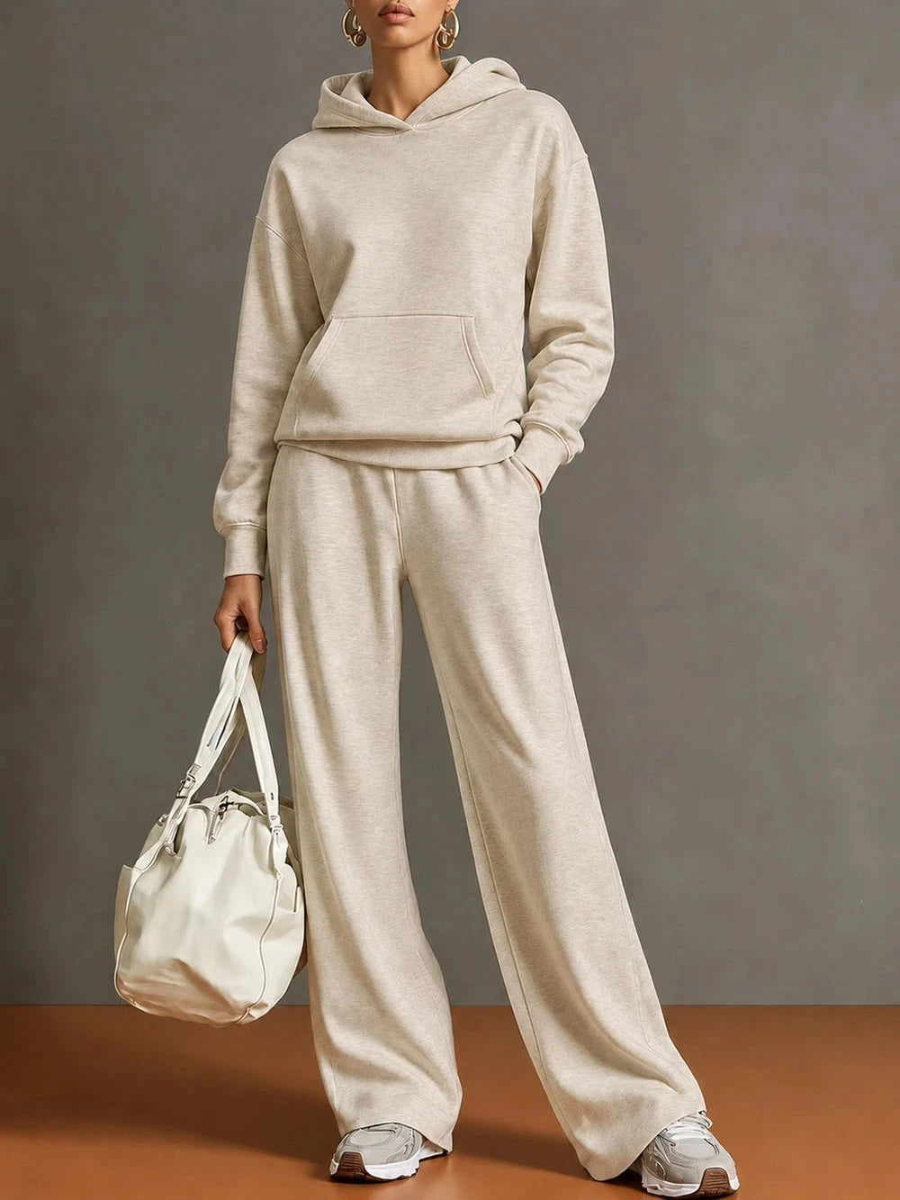 A model wearing a matching beige hoodie and wide-leg sweatpants set, holding a cream-colored bucket bag against a neutral gray studio background.