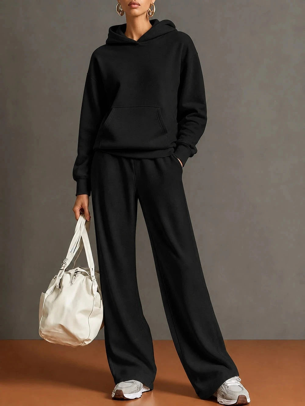 A model wearing a black pullover hoodie and matching black wide-leg sweatpants, holding a white shoulder bag against a gray studio backdrop.