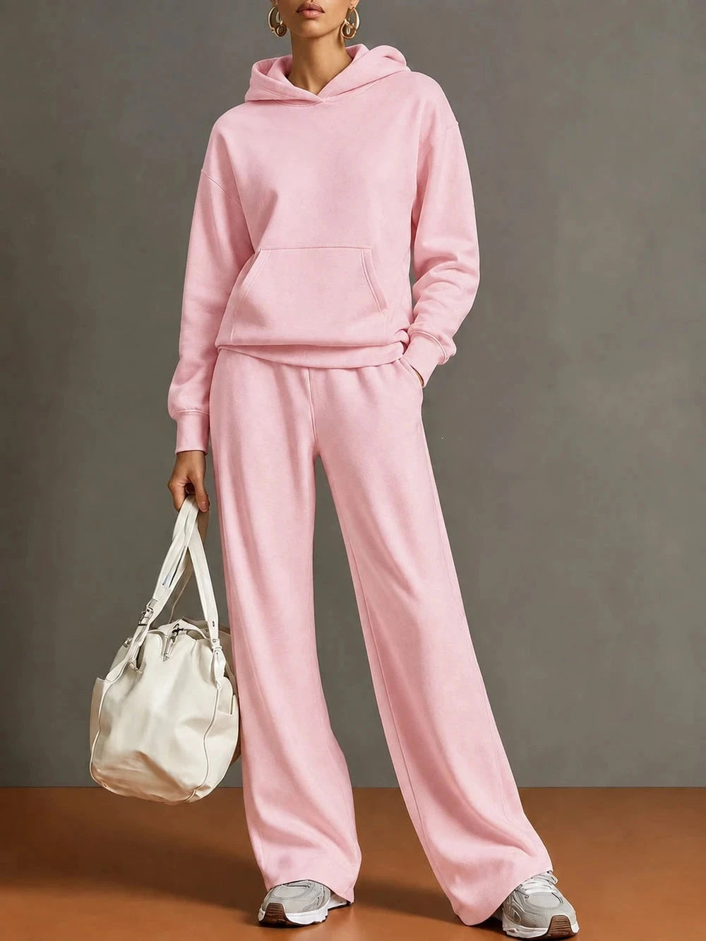 A model wearing a matching light pink two-piece loungewear set consisting of a pullover hoodie and wide-leg sweatpants, holding a cream-colored shoulder bag against a neutral gray studio background.