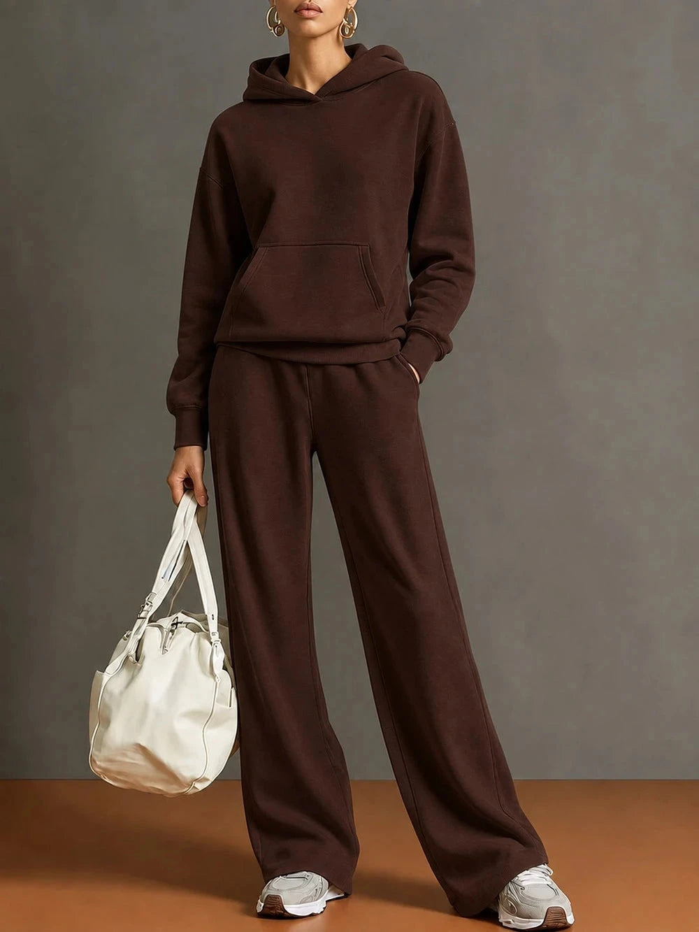 A model wearing a matching dark brown hoodie and wide-leg sweatpants set, holding a cream-colored leather tote bag against a neutral gray studio background.