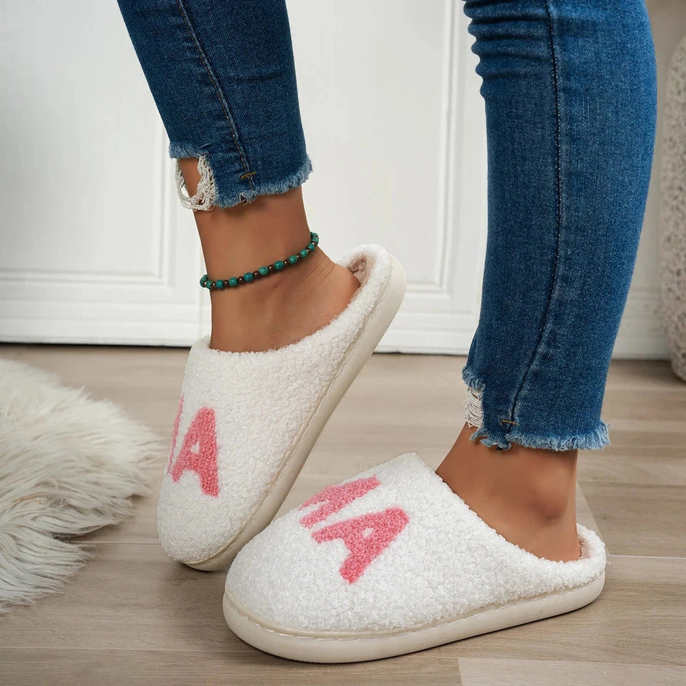 White slippers with pink letters worn by a person in blue jeans on a wooden floor.