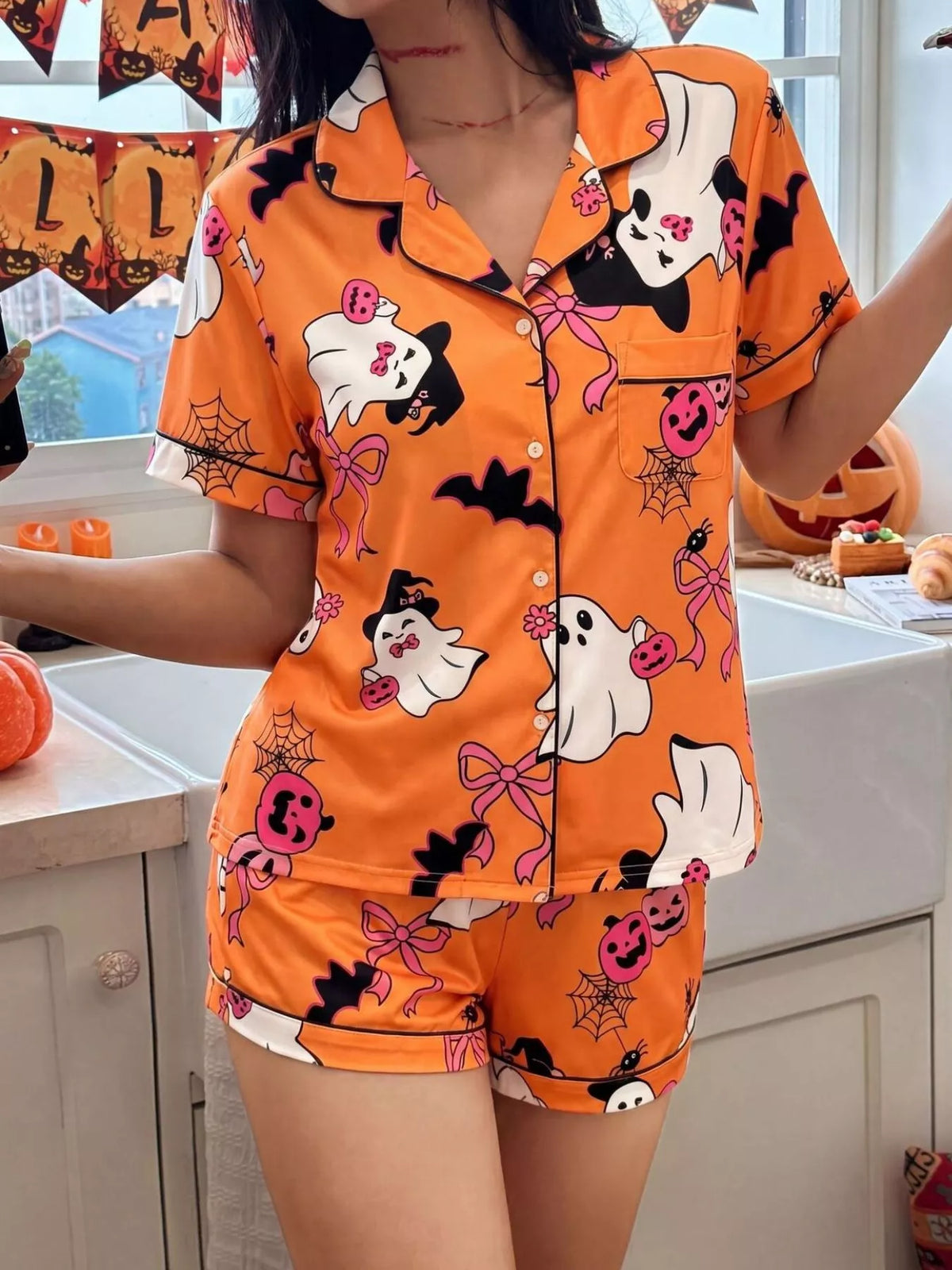 A woman wearing a two-piece orange pajama set with a Halloween print featuring ghosts, pandas, bats, and jack-o'-lanterns, standing in a kitchen setting decorated for the holiday.