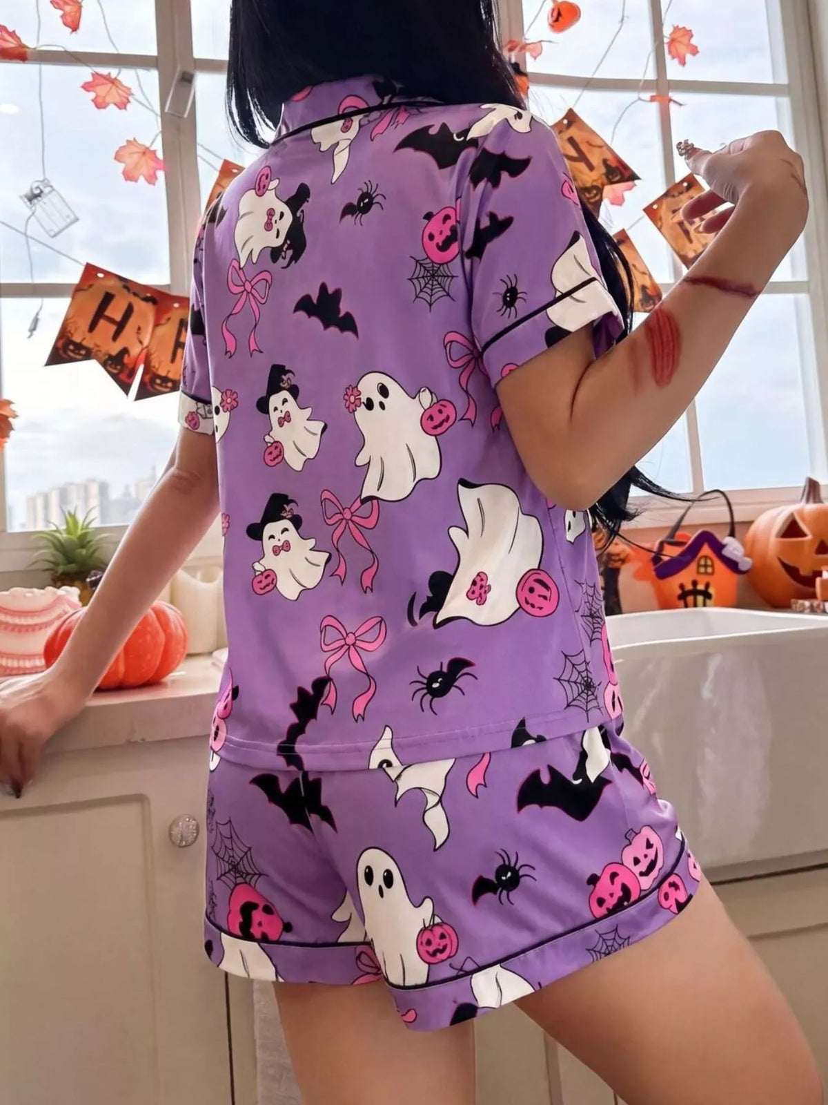 A person wearing a purple short-sleeve pajama set with a Halloween print of ghosts, bats, spiders, and pumpkins, standing in a decorated room near a window.