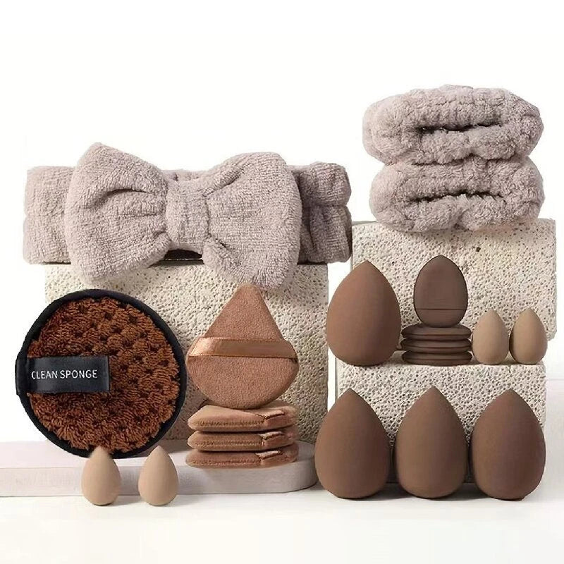 A coordinated set of makeup and skincare beauty tools arranged on stacked stone blocks against a clean white background. The set includes a large bow-shaped towel, two folded textured towels, a round sponge with a black label reading 'CLEAN SPONGE,' several teardrop-shaped makeup sponges, smaller sponge pieces, and multiple makeup blending sponges in earthy beige, taupe, and brown tones.