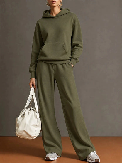 A model wearing an olive green matching lounge set consisting of a pullover hoodie and wide-leg sweatpants, holding a white tote bag against a neutral studio backdrop.