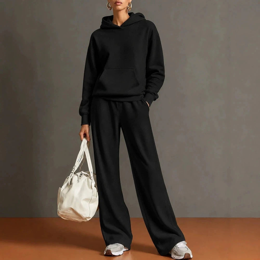 A model wearing a matching black hoodie and wide-leg sweatpants set, holding a white duffel bag against a gray studio backdrop with a warm brown floor.