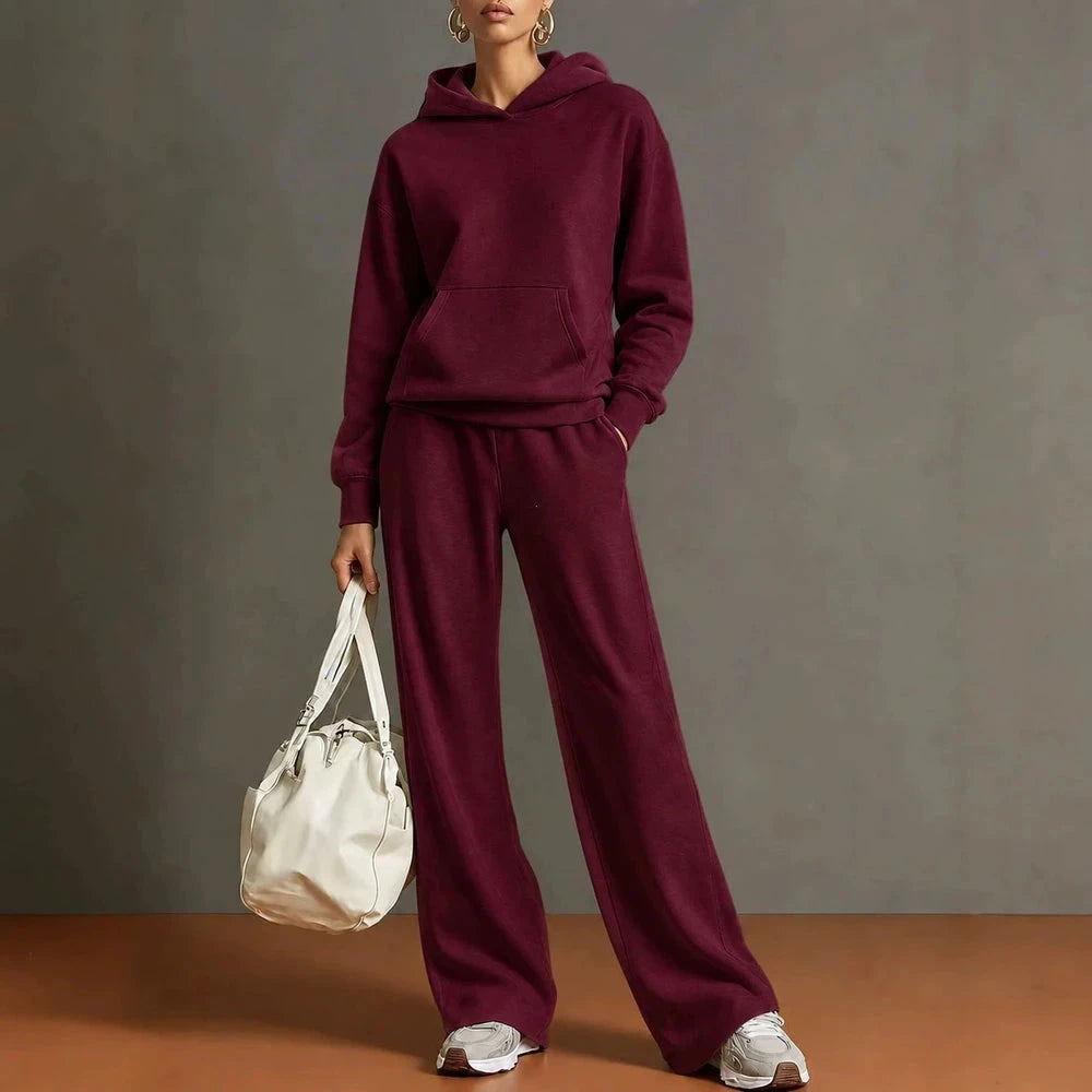 A model wearing a matching burgundy two-piece loungewear set consisting of a pullover hoodie and wide-leg sweatpants, holding a white handbag against a textured gray studio background.