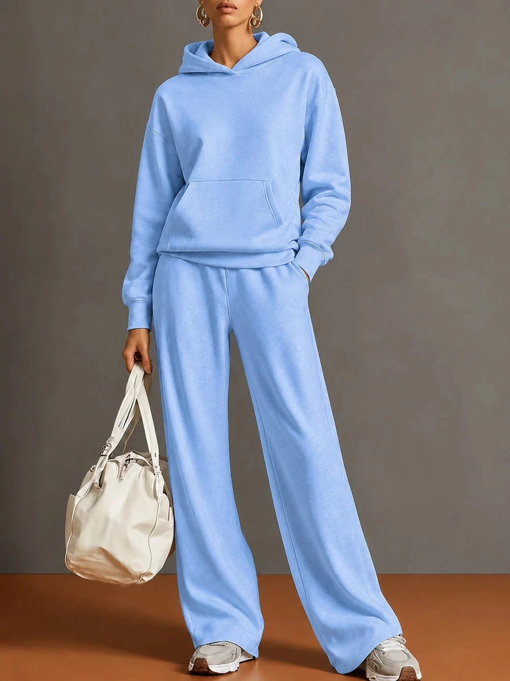 A model wearing a light blue matching hoodie and wide-leg sweatpants set, holding a cream-colored bucket bag, set against a neutral gray studio background.