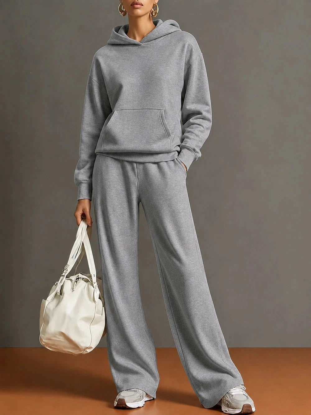 A model wearing a matching light gray two-piece loungewear set consisting of a pullover hoodie and wide-leg sweatpants, holding a cream-colored shoulder bag against a neutral gray studio background.