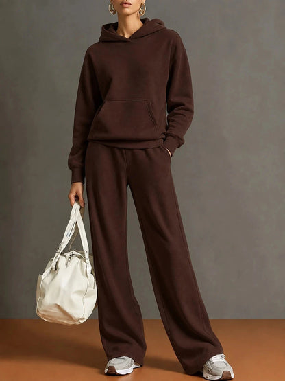 A model wearing a matching dark brown hoodie and wide-leg sweatpants set, holding a cream-colored leather tote bag against a neutral gray studio background.