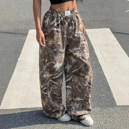 Model wearing loose wide-leg pants with a brown and green woodland camouflage print and elastic waistband, styled with white sneakers.