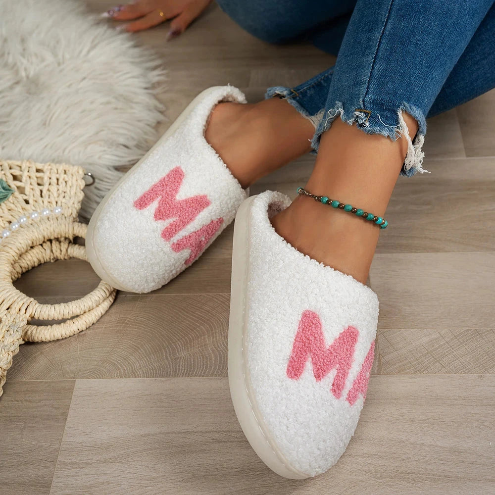 White slippers with pink letters worn by a person on a wooden floor.