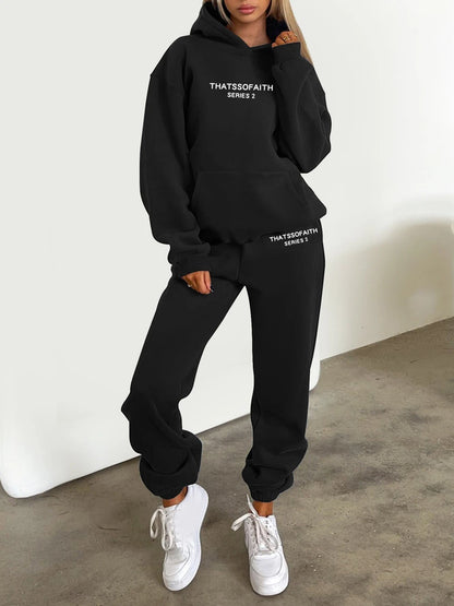Person wearing a black hoodie and pants with text, standing against a white wall.