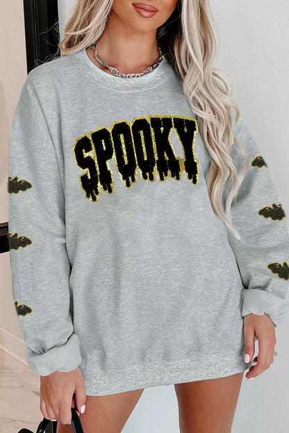 Woman modeling a gray Halloween crewneck sweatshirt with black dripping “SPOOKY” lettering outlined in gold and matching bat graphics on both sleeves.