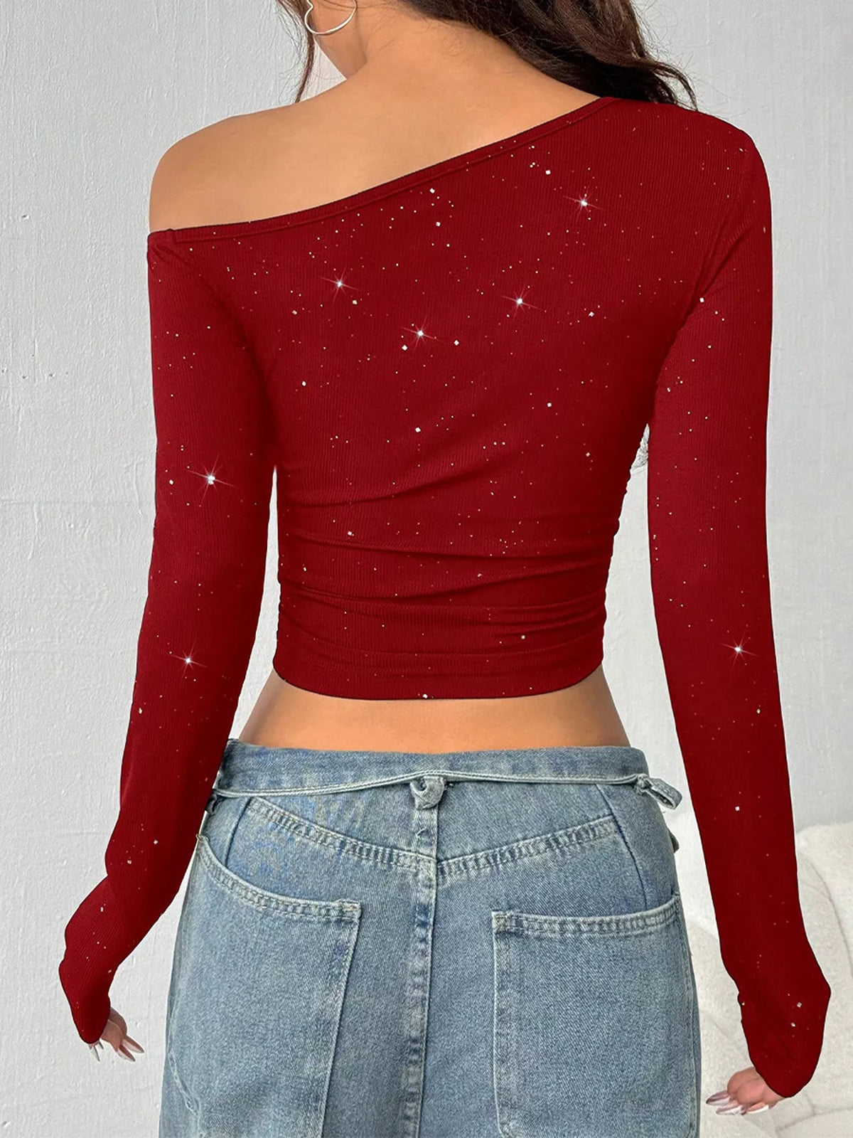 A woman shown from the back wearing a long-sleeve off-the-shoulder red crop top with subtle silver glitter speckles, paired with light blue jeans.