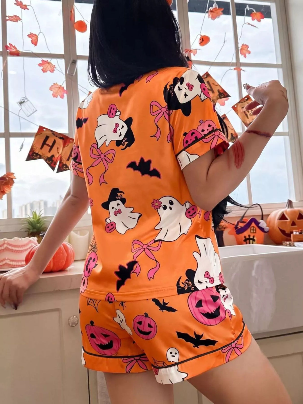 A woman wearing a bright orange Halloween-themed pajama set with a cartoon ghost, pumpkin, bat, and bow print, standing in a decorated room by a window.