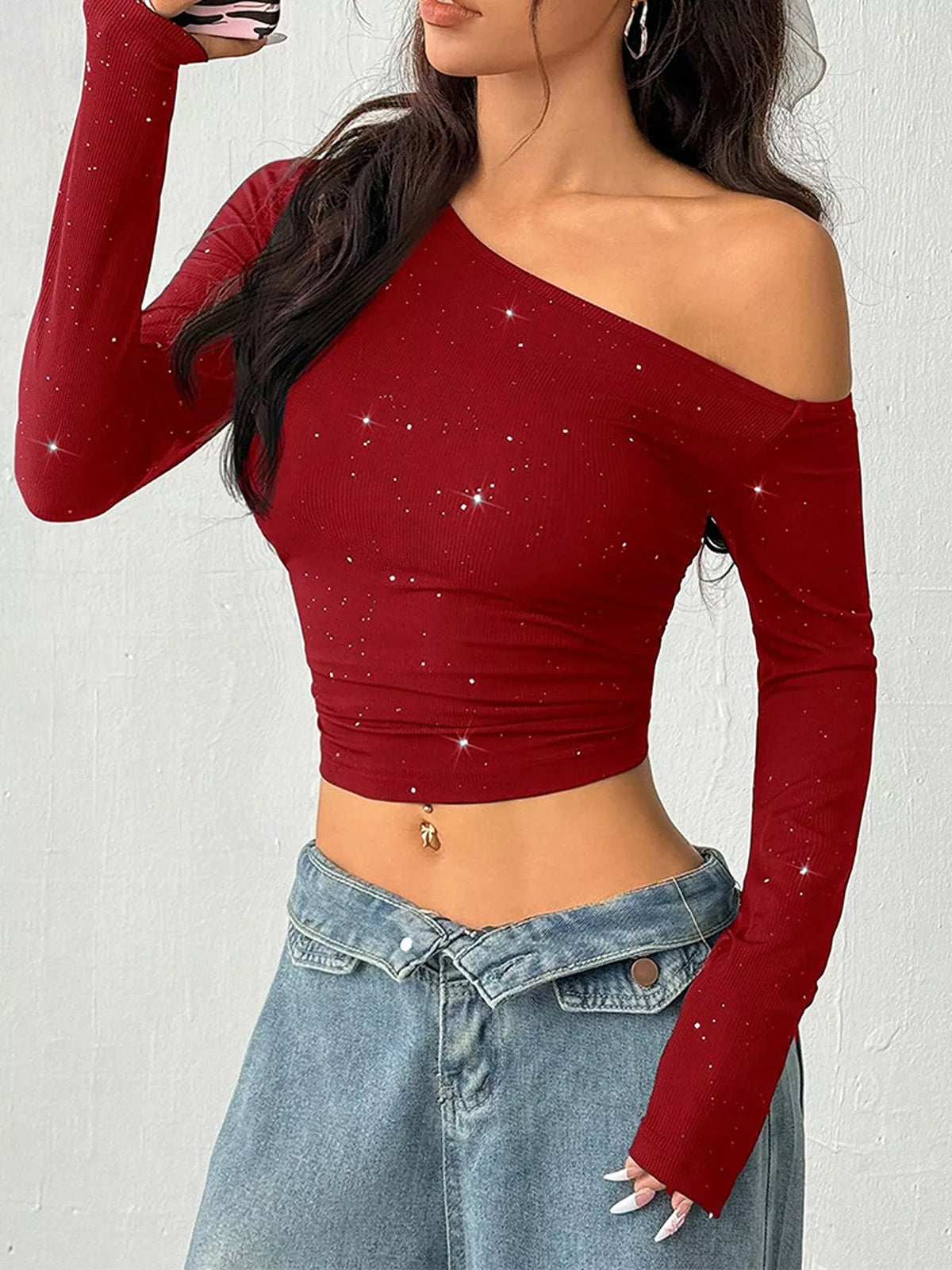 A woman wearing a long-sleeve off-the-shoulder crop top in deep red with a subtle sparkly pattern, paired with light blue jeans.