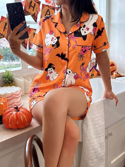 A woman takes a mirror selfie wearing a bright orange Halloween-themed pajama set with cartoon ghosts, pumpkins, bats, and spiderwebs. The setting is a kitchen with autumn decorations like pumpkins and a 'Halloween' banner.