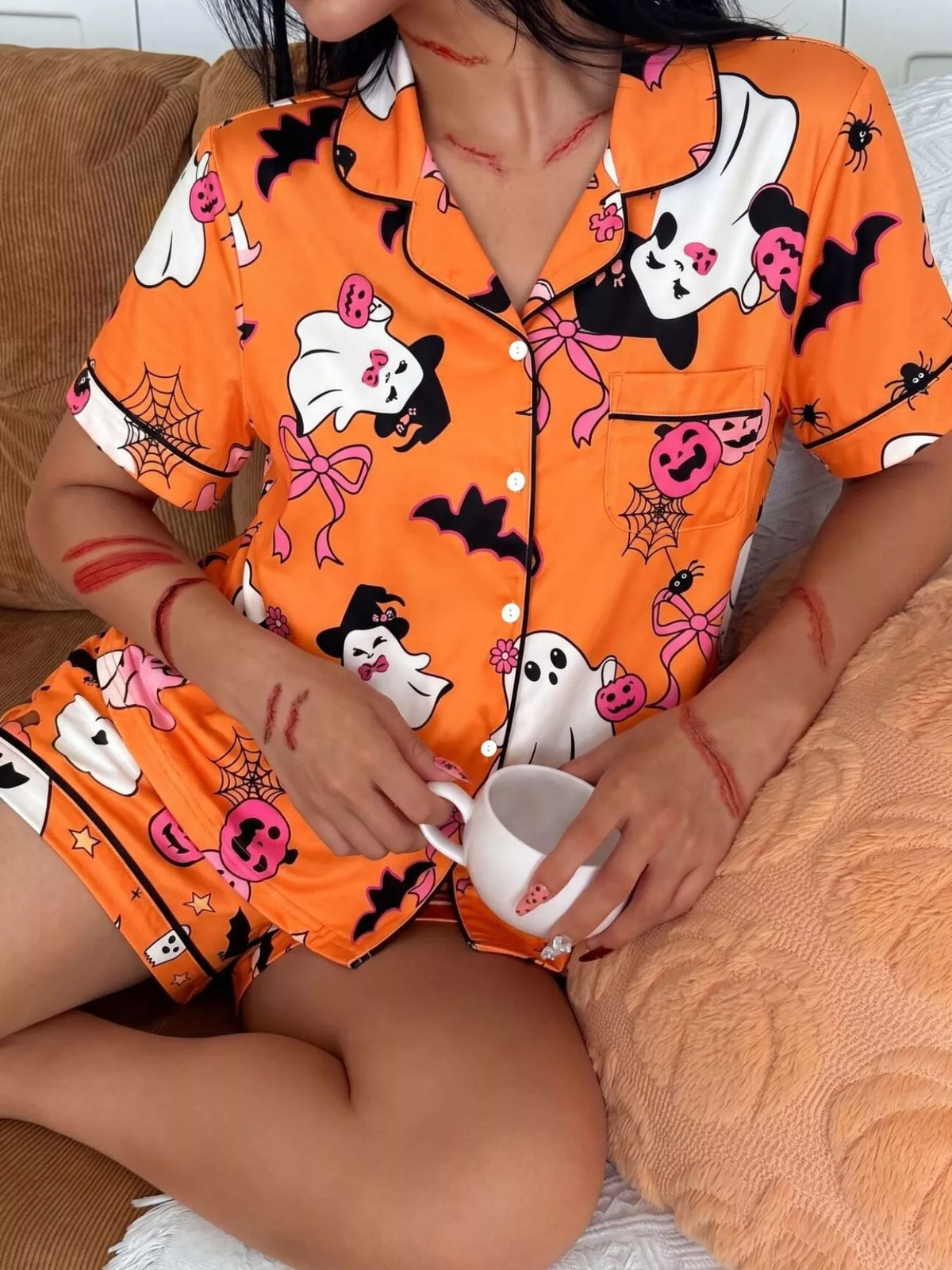 A person wearing a bright orange Halloween-themed pajama set with a cartoon ghost, spider, pumpkin, and bat print, sitting on a couch and holding a white mug.