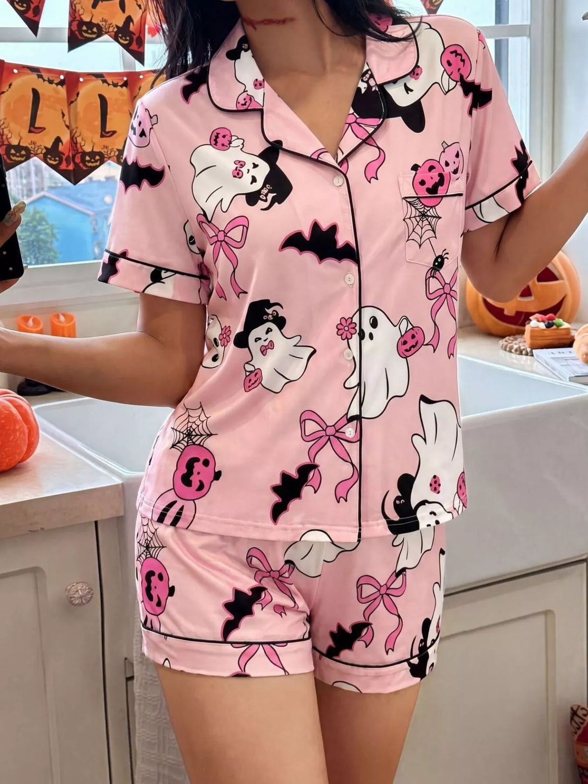 A woman wearing a pink short-sleeve pajama set with a Halloween print featuring ghosts, bats, pumpkins,  photographed in a kitchen setting.