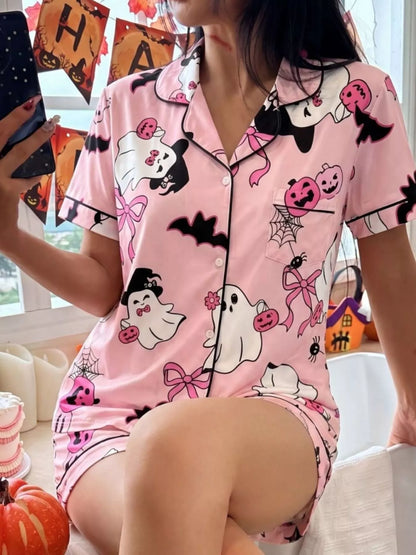 A woman wearing a pink short-sleeve pajama set with a Halloween print featuring cartoon ghosts, pumpkins, bats, and bows, photographed indoors near a window with holiday decorations.
