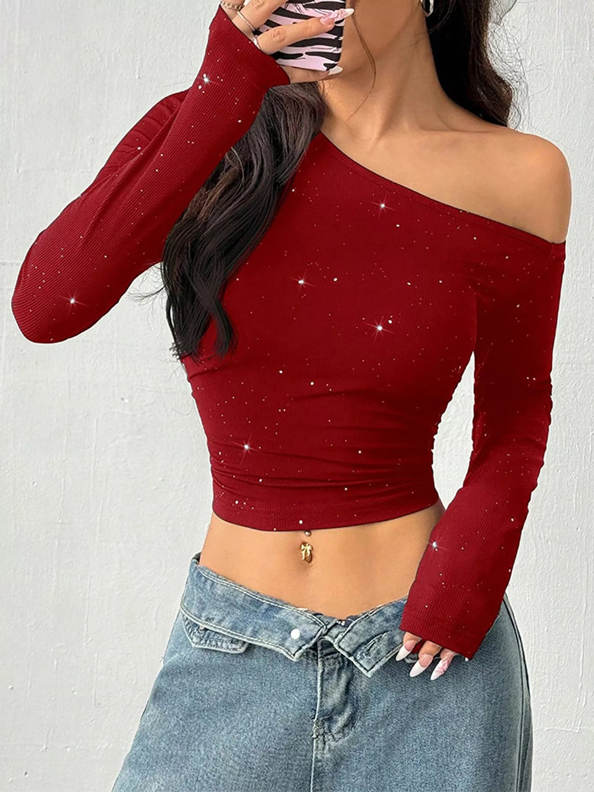 A woman wearing a long-sleeve off-the-shoulder crop top in deep red with subtle silver glitter speckles, paired with light blue denim jeans.