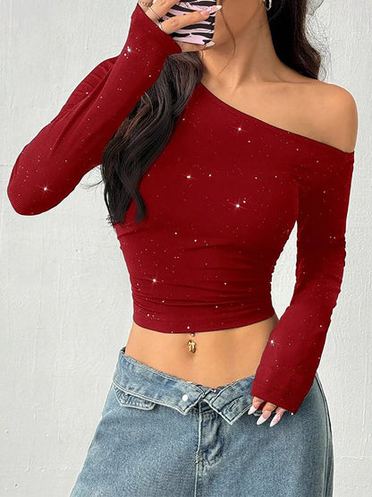 A woman wearing a long-sleeve off-the-shoulder crop top in deep red with subtle silver glitter speckles, paired with light blue denim jeans.