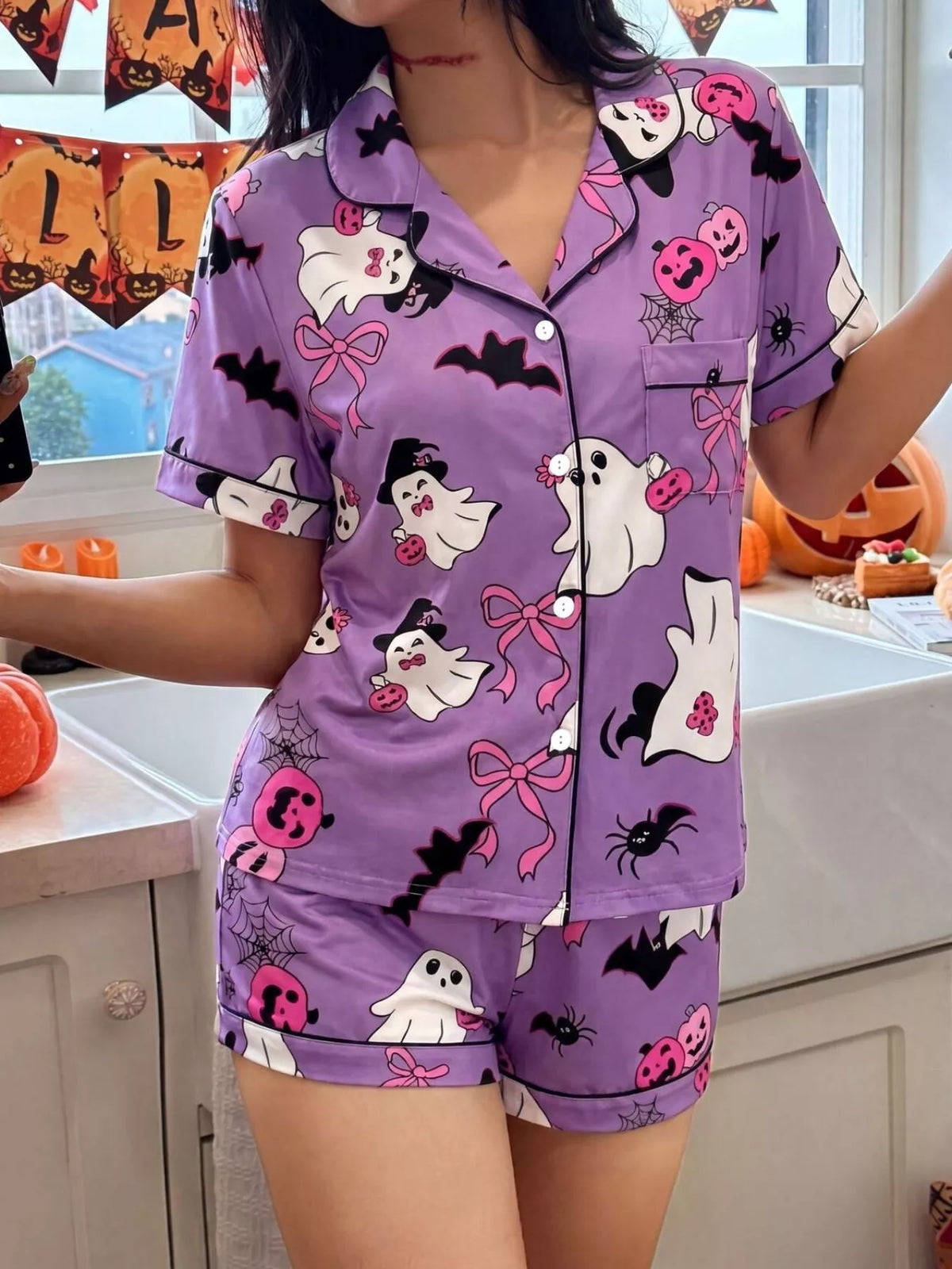 A woman wearing a purple short-sleeve pajama set with a Halloween print featuring ghosts, bats, spiders, and jack-o'-lanterns, standing in a kitchen decorated for Halloween.