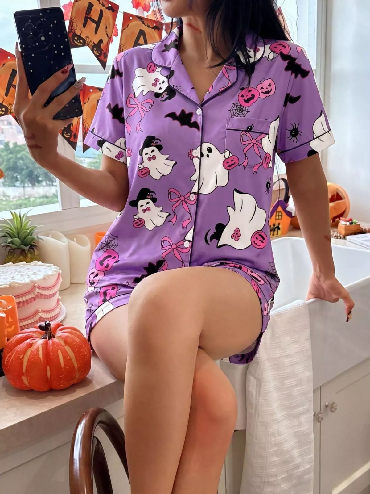 A woman takes a mirror selfie wearing a purple short-sleeve pajama set with a Halloween print featuring ghosts, pumpkins, bats, and bows. The setting is a brightly lit kitchen with autumn decorations.