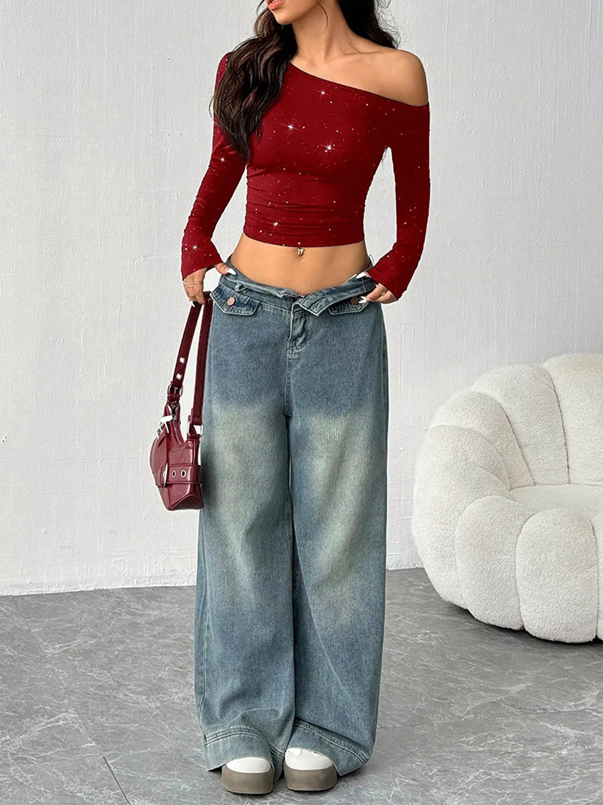 A woman models a red off-the-shoulder long-sleeve crop top with subtle sparkly details, paired with light-wash wide-leg jeans, holding a burgundy handbag.