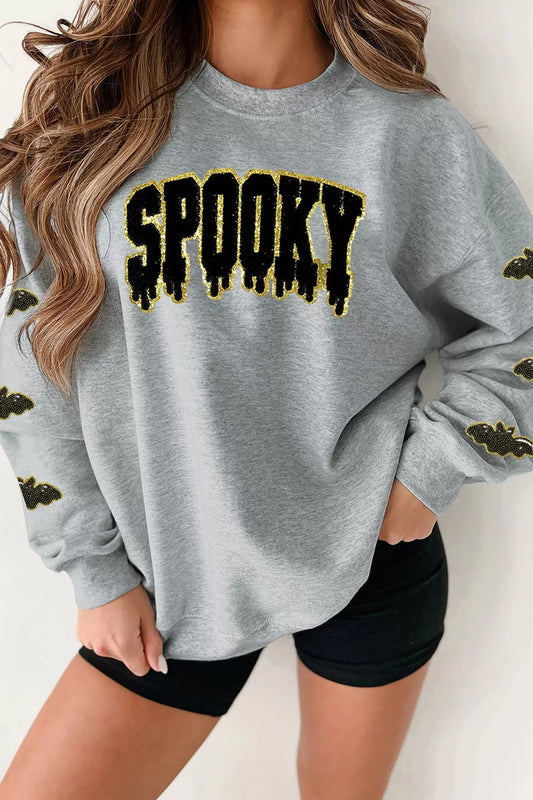 A woman wearing a light gray crewneck sweatshirt with 'SPOOKY' printed across the chest in black dripping letters outlined in yellow, paired with black shorts.